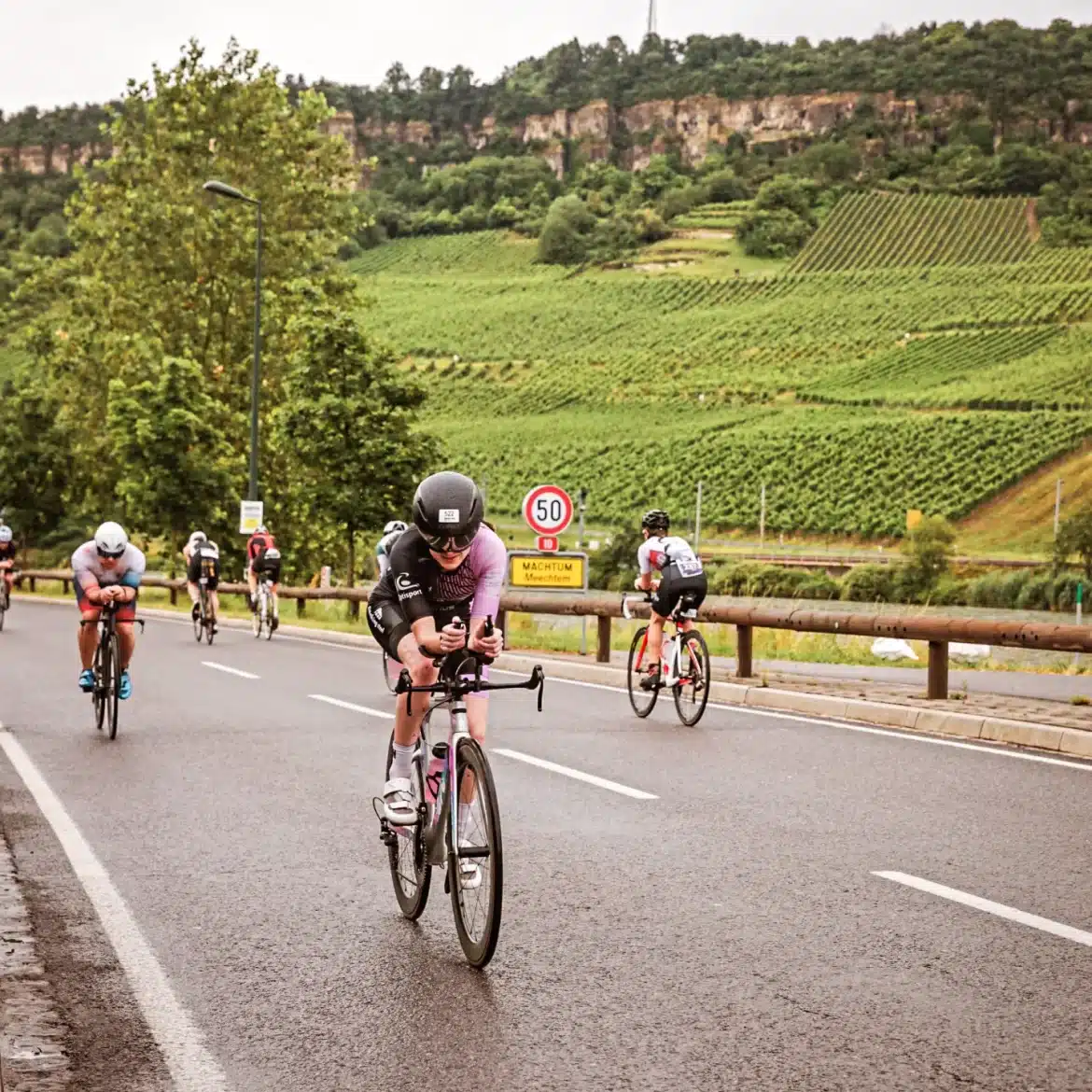 Female triathlete wearing a custom Pro 300 trisuit during a triathlon race in Machtem – purple-black aero suit on a time trial bike with hilly vineyards in the background
