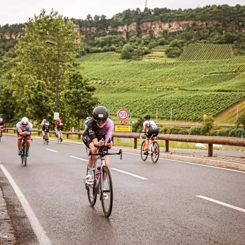 Female triathlete wearing a custom Pro 300 trisuit during a triathlon race in Machtem – purple-black aero suit on a time trial bike with hilly vineyards in the background
