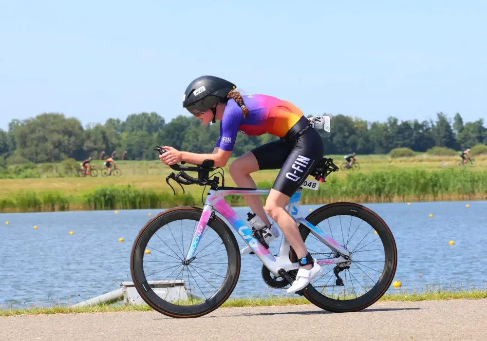 Woman wearing a custom Q-FIN Elite 1200 trisuit during triathlon – aerodynamic short sleeve suit in purple-orange gradient on a time trial bike by the water