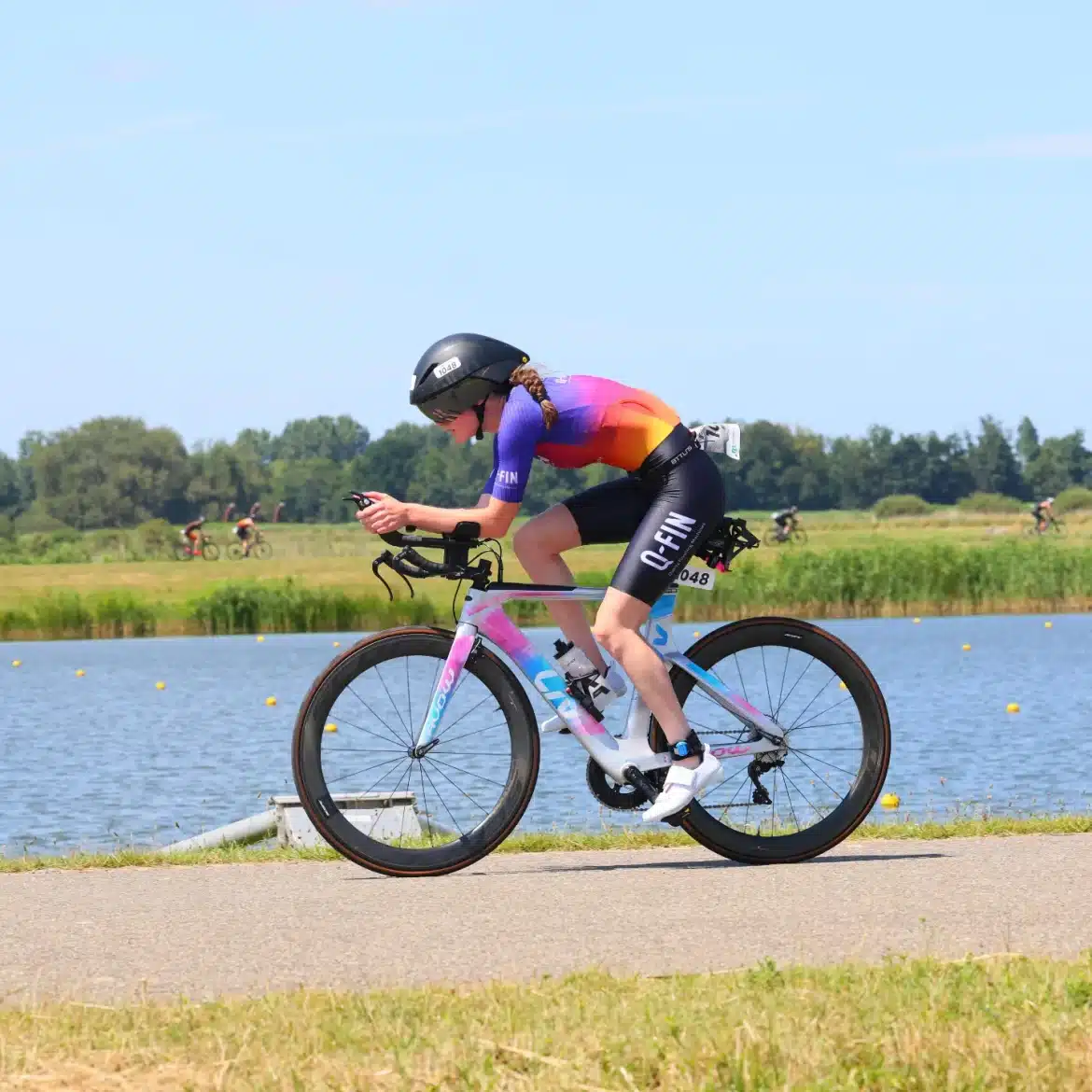 Woman wearing a custom Q-FIN Elite 1200 trisuit during triathlon – aerodynamic short sleeve suit in purple-orange gradient on a time trial bike by the water