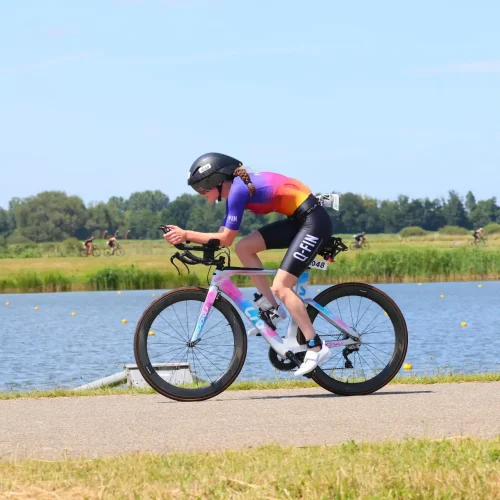Woman wearing a custom Q-FIN Elite 1200 trisuit during triathlon – aerodynamic short sleeve suit in purple-orange gradient on a time trial bike by the water